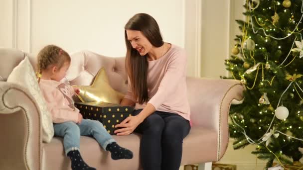 Mãe alegre e seu bebê bonito filha abrir um presente. Pais e criança se divertindo perto da árvore de Natal dentro de casa. Família amorosa Feliz Natal e Feliz Ano Novo . — Vídeo de Stock