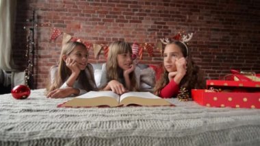 Üç kız kardeş yumuşak pijama yatağa yalan, üçüz Noel hikaye büyük bir kitap okumak, yatak odası Noel ışıkları ve hediyeler ile süslüdür