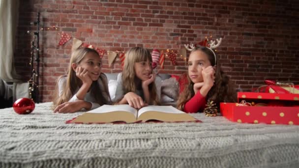 Três irmãs deitam-se na cama em pijamas macios, trigêmeos Leia um enorme livro de histórias de Natal, quarto é decorado com luzes de Natal e presentes — Vídeo de Stock