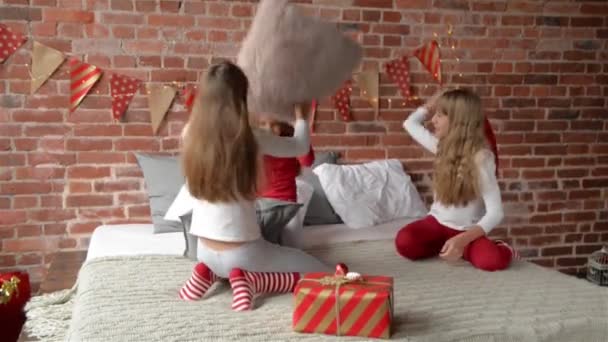 Las hermanas trillizas en pijama han arreglado la lucha por las almohadas. El dormitorio está decorado con guirnaldas de Navidad y regalos de Navidad. Trillizos niñas. Feliz Navidad y Feliz Año Nuevo . — Vídeos de Stock