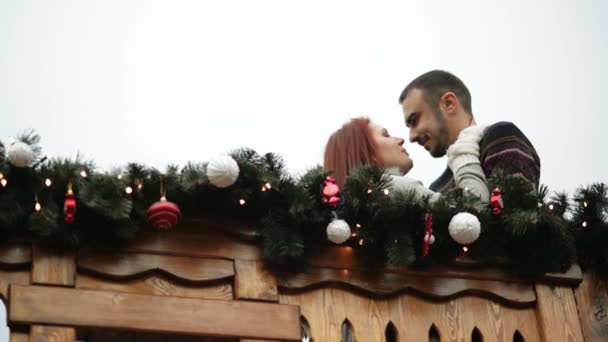 恋人のカップルは自分を抱きしめること。彼らはクリスマスの時間の間に木製のバルコニーに立っています。新年あけましておめでとうございますコンセプト. — ストック動画
