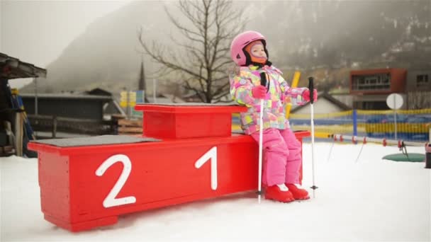 Πορτραίτο του μικρό χιονοδρομικό αρχάριος. Αυτή κάθεται σε ένα βάθρο νικητών. Χαριτωμένο παιδί έχει πολλή διασκέδαση δαπάνες χειμερινές διακοπές στο χιονοδρομικό κέντρο. — Αρχείο Βίντεο