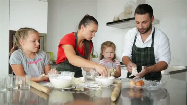 Las niñas lindas y sus hermosos padres amasan la masa y sonríen mientras cocinan en la cocina en casa . — Vídeo de stock