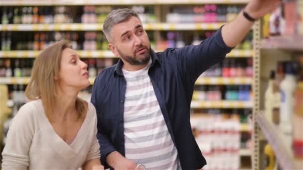 Feliz pareja de compras en el supermercado. Joven familia eligiendo vino de los estantes del supermercado . — Vídeo de stock