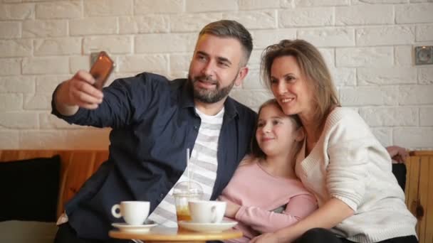 Glückliche Mutter, Vater und kleines Mädchen haben Spaß und machen Selfie mit dem Smartphone im Café. — Stockvideo