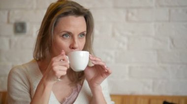 Genç güzel kadın Cafe ile fincan çay veya kahve ve Gulping içinde oturur. Mutlu kız içki içecek ve penceresine bakarak.