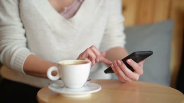 Frau am Tisch, Kaffee oder Tee trinkend. Sie rührt an einer Tasse und schaut aufs Smartphone. Nahaufnahme — Stockvideo
