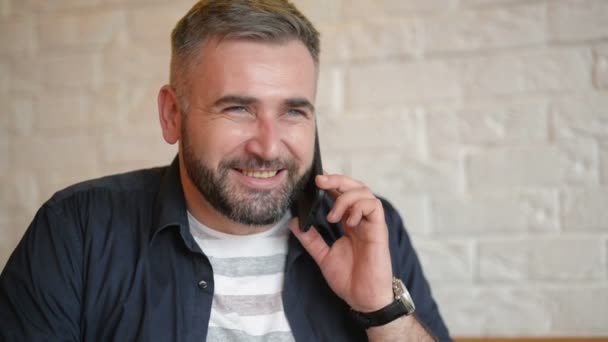 Rozmawia przez telefon w Cafe przystojny biznesmen. Młody Brodaty facet trzyma jego smartfon w pobliżu jego ucha. Atrakcyjny mężczyzna kaukaski w białej koszuli, nazywając kogoś na swój telefon komórkowy. — Wideo stockowe