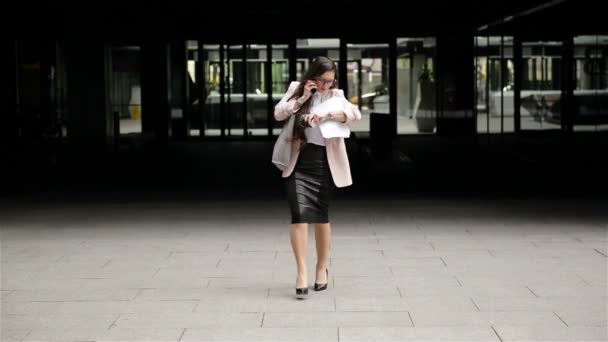 En ung stressande affärs kvinna i glasögonen går genom staden med dokument och samtal på telefonen. Hon droppar hennes papper för stress. — Stockvideo
