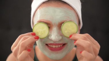 Bir kızın yüzünde bir maske ve gözleri gülümser ve salatalık ile kapatan başında bir havlu ile kapatın. Kadın giyen yüz maskesi. Yüz bakım kavramı