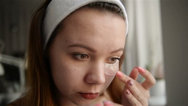 Het meisje in de buurt van de spiegel voert schoonheidsbehandelingen op het gezicht. Het meisje zet patches op het gebied onder de ogen. — Stockvideo