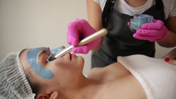 Cosmetologist teszi eljárás tisztító és hidratáló a bőr. Alkalmazása maszk különleges ecsettel, hogy szembenézzen a nő. Fiatal lány gondoskodik a bőre. Látogatás kozmetológiai Klinika. Aethetic orvostudomány. — Stock videók