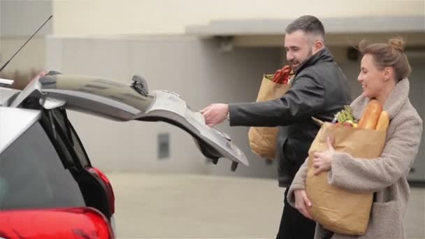 La joven familia está empacando sus bolsas de comestibles en un auto nuevo. Vista frontal del hombre barbudo guapo y la mujer hermosa . — Vídeos de Stock