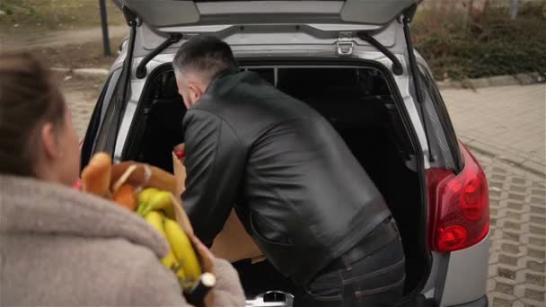 Taze yiyeceklerle dolu alışveriş arabası olan genç ve şık bir çift açık hava otoparkındaki arabaya eşyalarını dolduruyor.. — Stok video