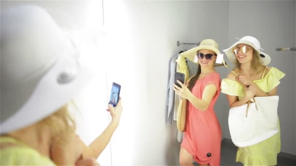 Mulher feliz tentando um novo óculos de sol no guarda-roupa na loja de roupas. Amigos femininos se divertindo roupas de compras. Senhoras fazendo espelho Selfie no quarto de montagem . — Vídeo de Stock