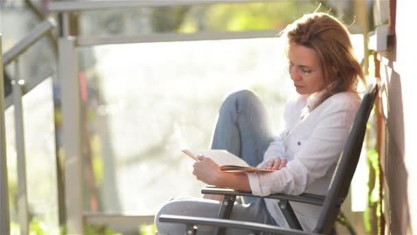 Porträt eines jungen schönen Mädchens mit kaukasischem Aussehen las interessantes Buch auf einem Balkon sitzend. Frau entspannt sich im Urlaub. — Stockvideo