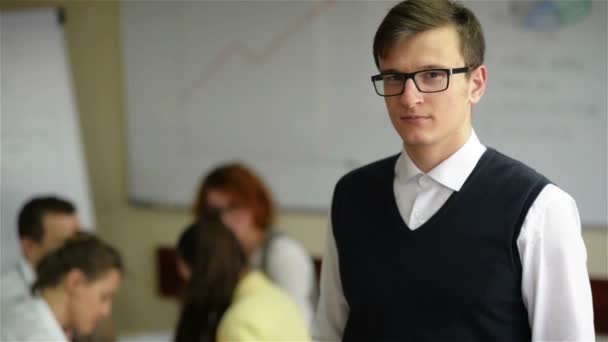 Gros plan d'un beau jeune travailleur portant des lunettes et une chemise en flanelle dans un petit bureau avec d'autres membres du personnel en arrière-plan — Video