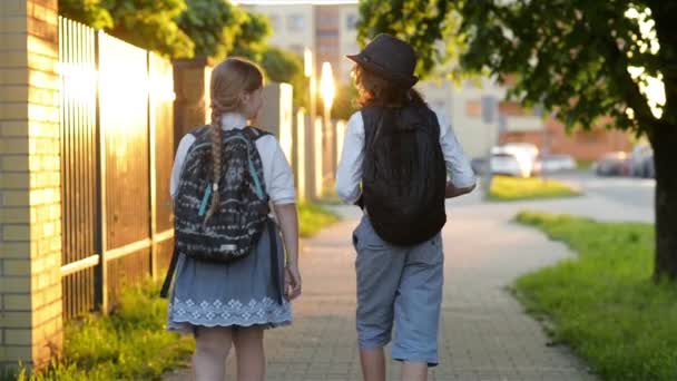 二人の小学生がバックパックを持って学校に行きます。かわいい子供ティーンガールとバッグを持つ男の子は学校に戻って。友人、幼年期と自己教育の概念. — ストック動画