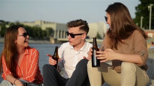 4人のスタイリッシュなハッピーヒップスターがビールのボトルを持って公園を歩き、一緒に抱き合っています。彼らはボトルでクリンクします。スローモーションビュー. — ストック動画