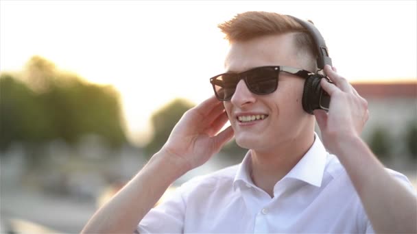 音楽を聴くハンサムな白人学生の肖像画。カメラを見てヘッドフォンとサングラスを持つ幸せな笑顔の男。クローズアップ, アウトドアコンセプト. — ストック動画