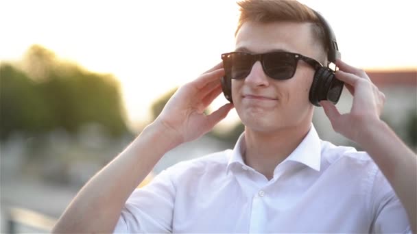Porträt eines gutaussehenden kaukasischen Studentenjungen, der Musik hört. Glücklich lächelnder Mann mit Kopfhörern und Sonnenbrille, der in die Kamera blickt. Nahaufnahme, Outdoor-Konzept. — Stockvideo