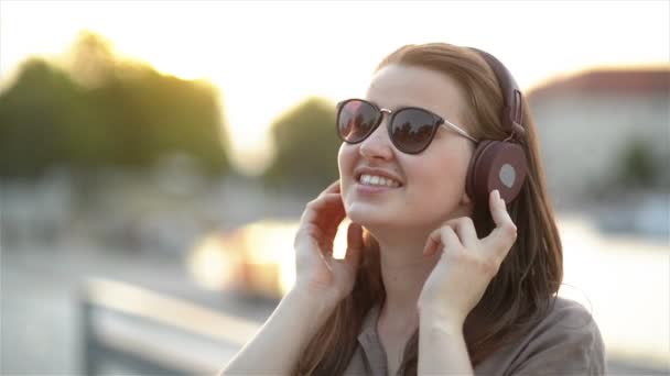 Gros plan Portrait drôle de jeune jolie fille écoutant de la musique sur les gros écouteurs, portant des lunettes de soleil mignonnes, posant au centre-ville au jour ensoleillé d'été . — Video