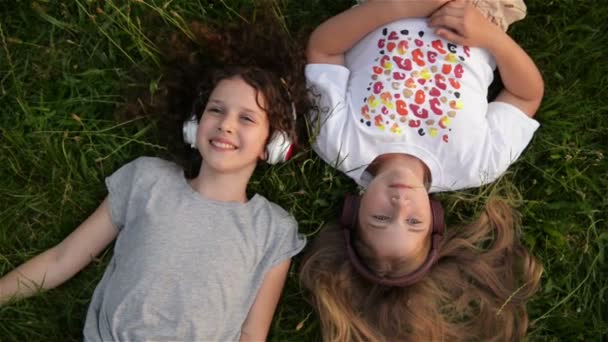 Dos chicas lindas usando auriculares acostadas en el parque. Hermosos niños en edad escolar se divierten pasando tiempo juntos . — Vídeos de Stock