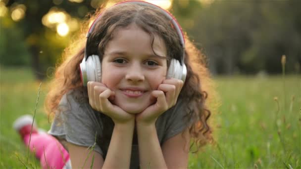 Dívka ležící na trávě a naslouchající hudbě ve sluchátkách. Rozkošný malý Kavkazský kluk se baví venku venku. — Stock video