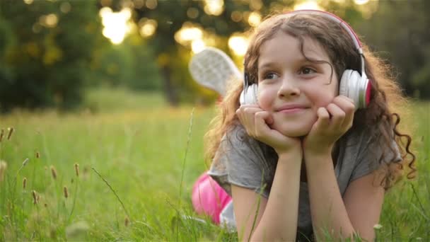 Κορίτσι ξαπλωμένη στο γρασίδι και ακούγοντας μουσική στα ακουστικά. Χαριτωμένο μικρό καυκάσιο παιδί έχει τη διασκέδαση ψύξη έξω σε εξωτερικούς χώρους. — Αρχείο Βίντεο