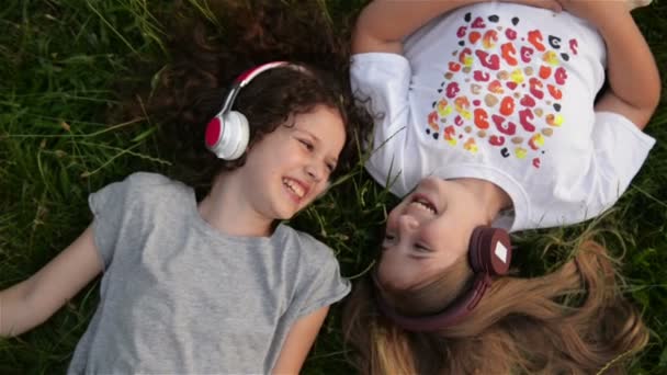 Dos chicas lindas usando auriculares acostadas en el parque. Hermosos niños en edad escolar se divierten pasando tiempo juntos . — Vídeos de Stock