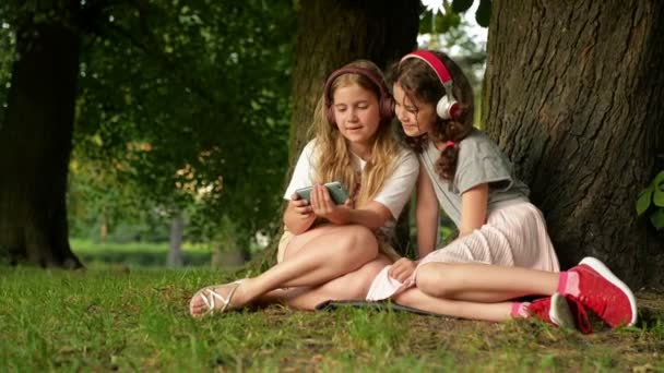 Dos hermanas jóvenes lindas con auriculares que se divierten en hermoso día. Niños felices jugando en el parque de verano. Niños viendo algo interesante por teléfono. Actividades de verano para niños . — Vídeos de Stock