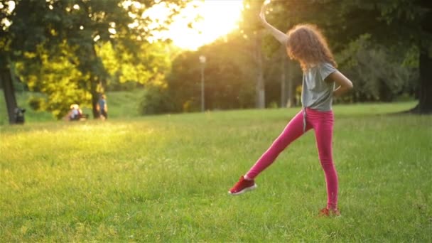 ドレスの幸せな小さな女の子は、公園の牧草地にジャンプし、アクロバットホイールを作っています。夏のアウトドアアクティビティ。スローモーション. — ストック動画