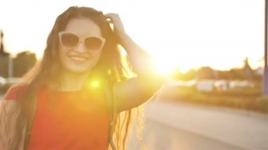 Kıvırcık saçlı güzel genç kadının geri görünümü şehir caddesinde yürürken, sonra kameraya döner, neşeyle gülümseyen Anlar mutluluk neşeli ruh hali. Yavaş çekim, kadın portresi, güneş merceği