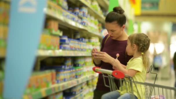 Menina bonito com aparência caucasiana está comendo pirulito e sentado em carrinho de supermercado. Mãe no fundo escolhe comida de bebê para sua filha . — Vídeo de Stock