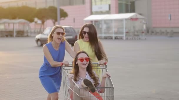 Amigos entretenidos en el estacionamiento del centro comercial en el día brillante. Las mujeres enérgicas rodeando atractiva chica alegre en el carrito de compras disfrutando del tiempo libre, slomo — Vídeo de stock