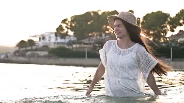 Девушка в платье брызгает бирюзовой водой руками, когда она стоит в океане и вращается из стороны в сторону. Женщина-туристка наслаждается летними каникулами . — стоковое видео