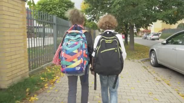 Dos amigos con mochilas van a la escuela en las calles de la ciudad más allá de los coches. Hermana con su hermano de vuelta a la escuela. El sol está en el fondo . — Vídeos de Stock