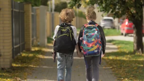 Twee Kind Meisje en Jongen wandelen door de straten langs auto 's. Zuster met haar broer terug naar school. De zon staat op Achtergrond. Achteraanzicht — Stockvideo