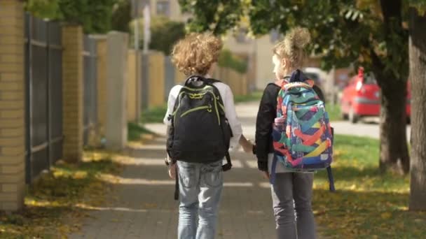 Δύο κορίτσια και ένα αγόρι περπατούν στους δρόμους της πόλης μπροστά από τα αυτοκίνητα. Αδελφή με τον αδελφό της πίσω στο σχολείο. Παιδιά μεταφέρουν σακίδια, πίσω όψη — Αρχείο Βίντεο