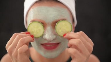 Bir kızın yüzünde bir maske ve gözleri gülümser ve salatalık ile kapatan başında bir havlu ile kapatın. Kadın giyen yüz maskesi. Yüz bakım kavramı