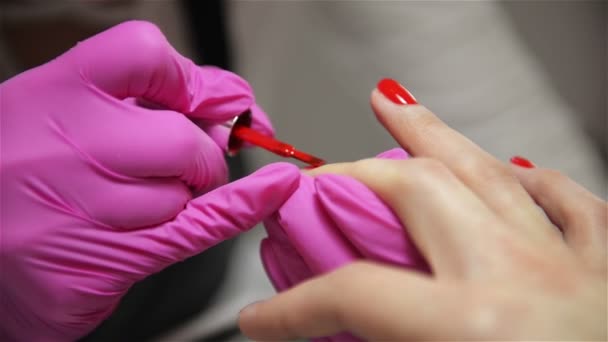 Femme ayant une manucure d'ongles dans un salon de beauté avec une vue rapprochée d'un esthéticienne appliquant un vernis avec un applicateur. Maître ongles peints avec vernis à ongles. Détails des mains appliquant l'ongle rouge . — Video