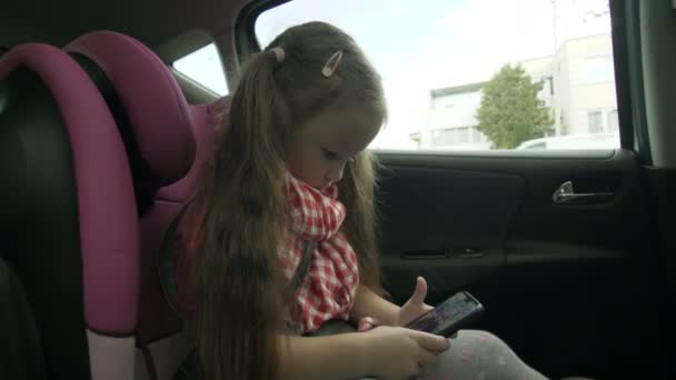 Χαριτωμένο κοριτσάκι απασχολημένος με έξυπνο τηλέφωνο, ενώ κάθεται στο πίσω κάθισμα του αυτοκινήτου. Υπέροχο παιδί στοιχειώδη ηλικία παίζει στο κινητό τηλέφωνο κατά τη διάρκεια του ταξιδιού — Αρχείο Βίντεο