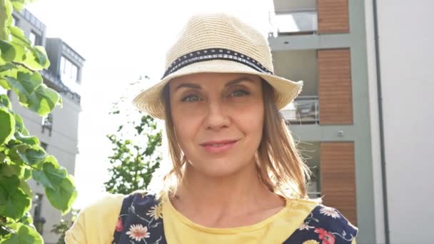 Portrait of a smiling woman in a straw hat. She stands in the courtyard of an apartment building. Spring, sun, good mood. — Stock Video