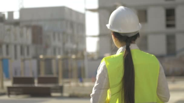 Kobieta w kamizelce sygnalizacyjnej i twardym kapeluszu na tle obiektu w budowie. Widok z tyłu. — Wideo stockowe