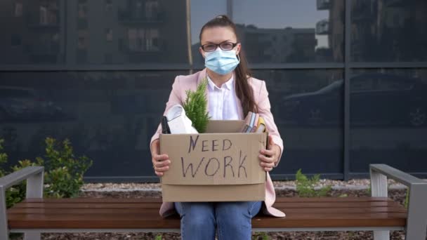 办公室旁边的长椅上坐着一位戴着医疗面具的悲伤的年轻妇女。她手里拿着一个装有个人物品的盒子。盒子上写着"需要工作" 。在Covid-19大流行病期间失业人员. — 图库视频影像