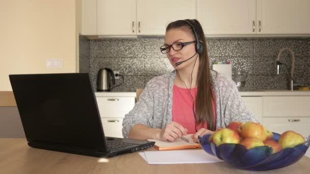 Młoda kobieta z zestawem słuchawkowym przed laptopem przy stole, konsultacja online z klientem. — Wideo stockowe