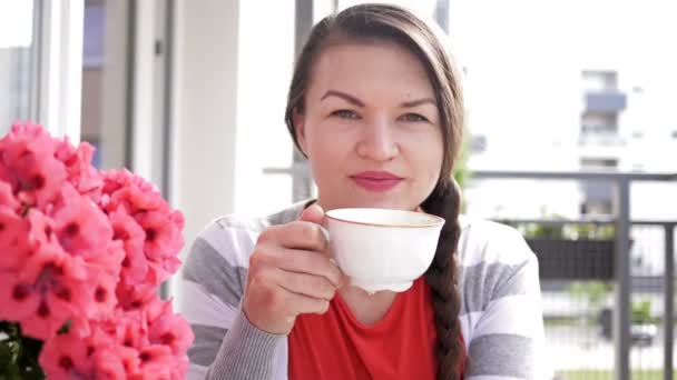 Retrato de una guapa morena con una taza de café de la mañana. Mujer está sentada en el balcón y sonriendo felizmente . — Vídeos de Stock