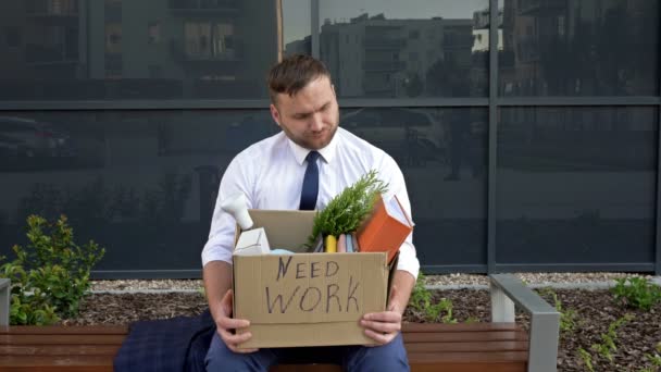 Sur le banc près du bureau se trouve un homme d'âge moyen très contrarié. Dans ses mains se trouve une boîte avec des effets personnels. Sur la boîte il est dit BESOIN DE TRAVAIL. — Video