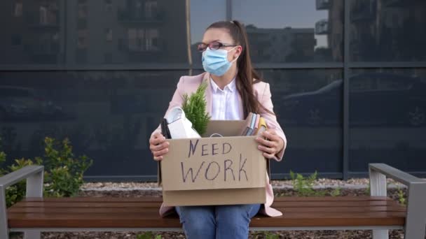 On the bench near the office sits a sad young woman in a medical mask. In her hands is a box with personal belongings. On the box it says NEED WORK. During the Covid-19 pandemic. Unemployed. — Stock Video