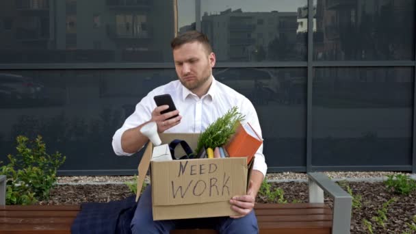 Precisam de trabalho. No banco perto do escritório fica um homem de meia-idade muito chateado. Em suas mãos está uma caixa com pertences pessoais e telefone celular. Homem está tentando encontrar trabalho usando a Internet . — Vídeo de Stock